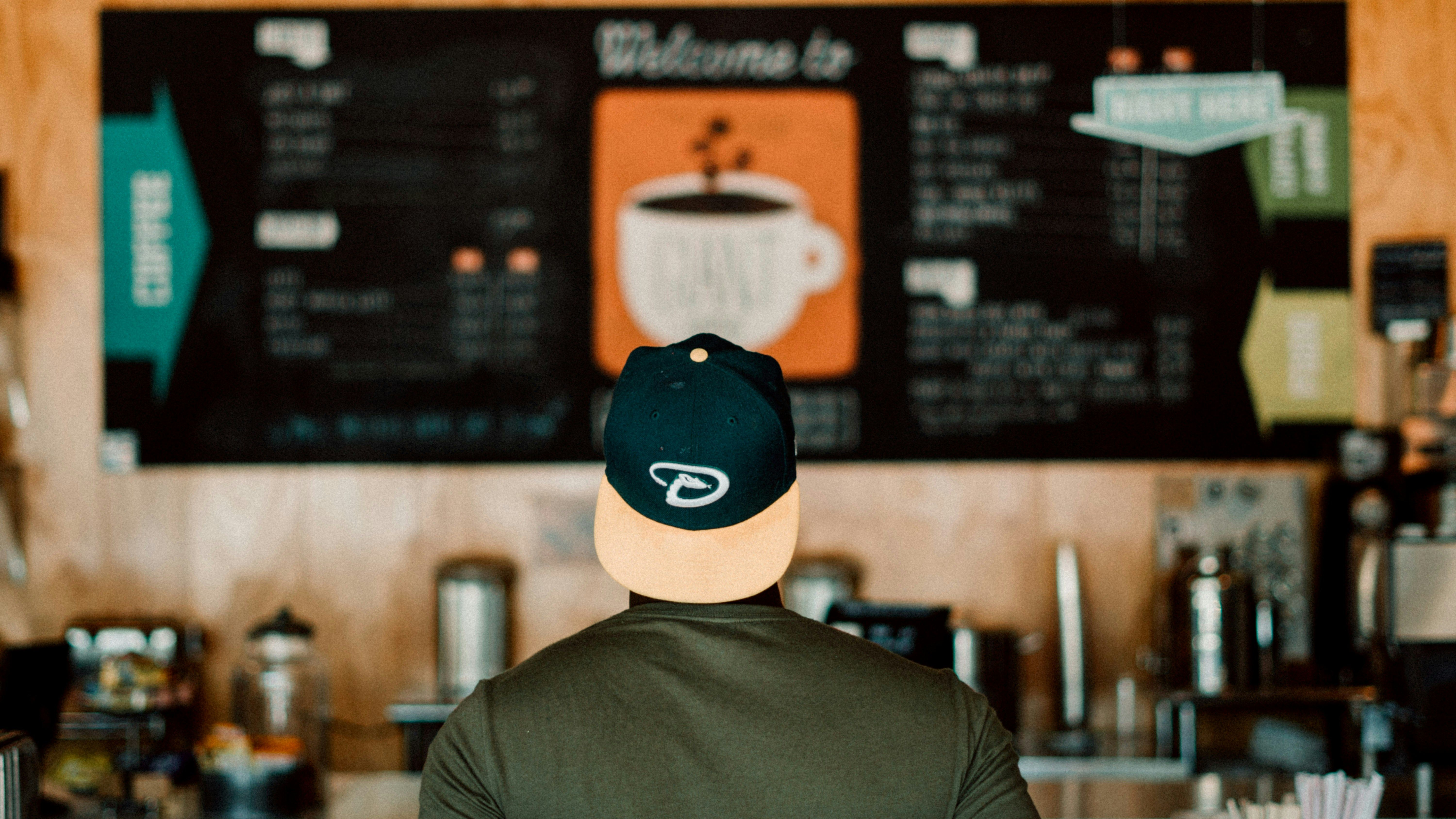 un ragazzo con un cappello è di spalle e osserva il menù di una caffetteria